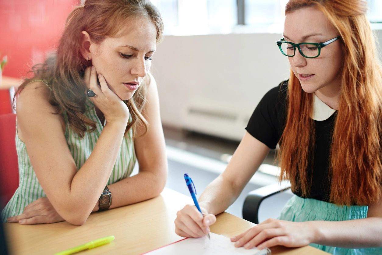 Two women sitting at a table writing on paper.