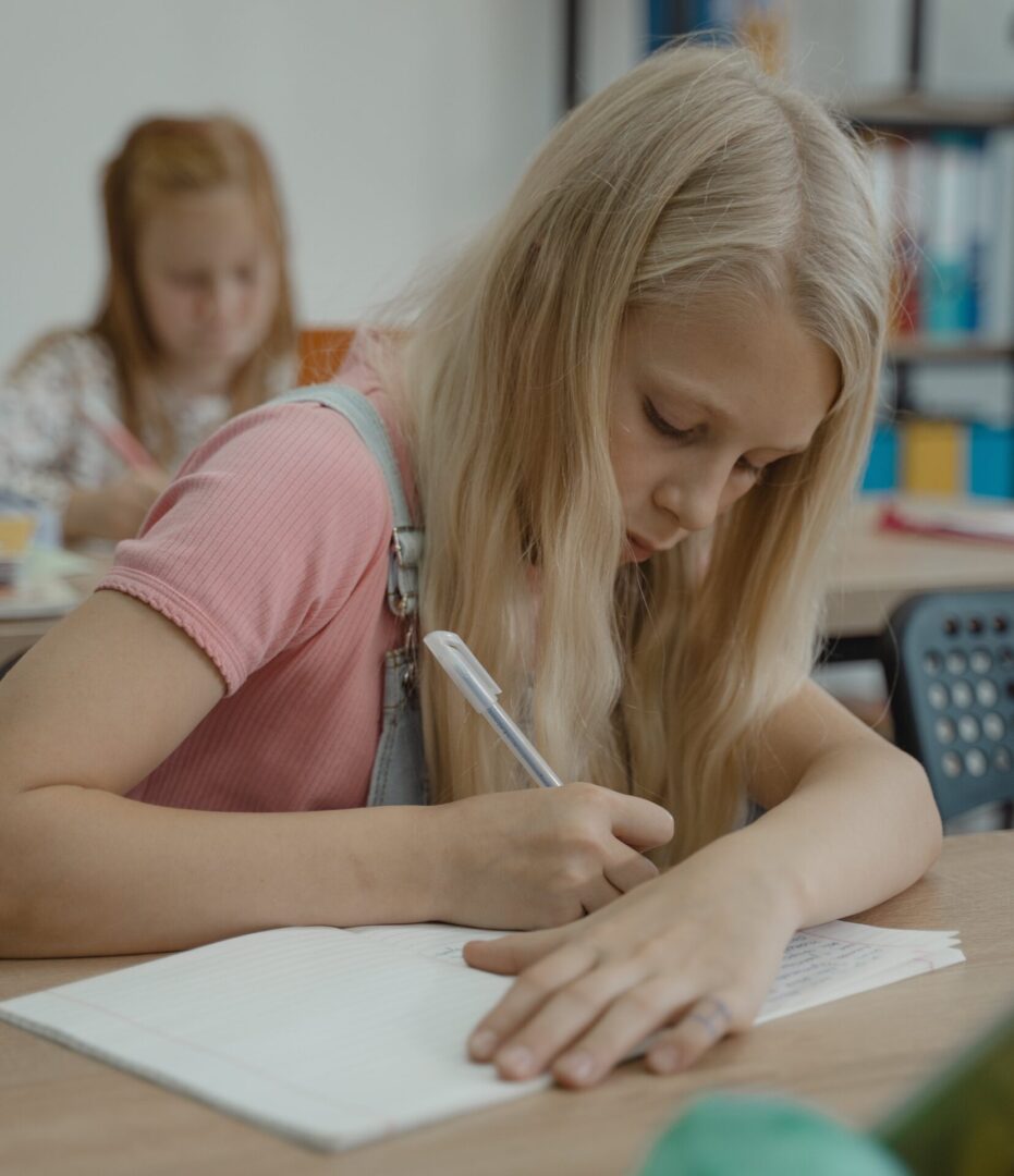 A girl writing on paper with another girl sitting in the background.